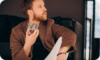 Man sitting indoors, holding a glass, with a thoughtful expression.