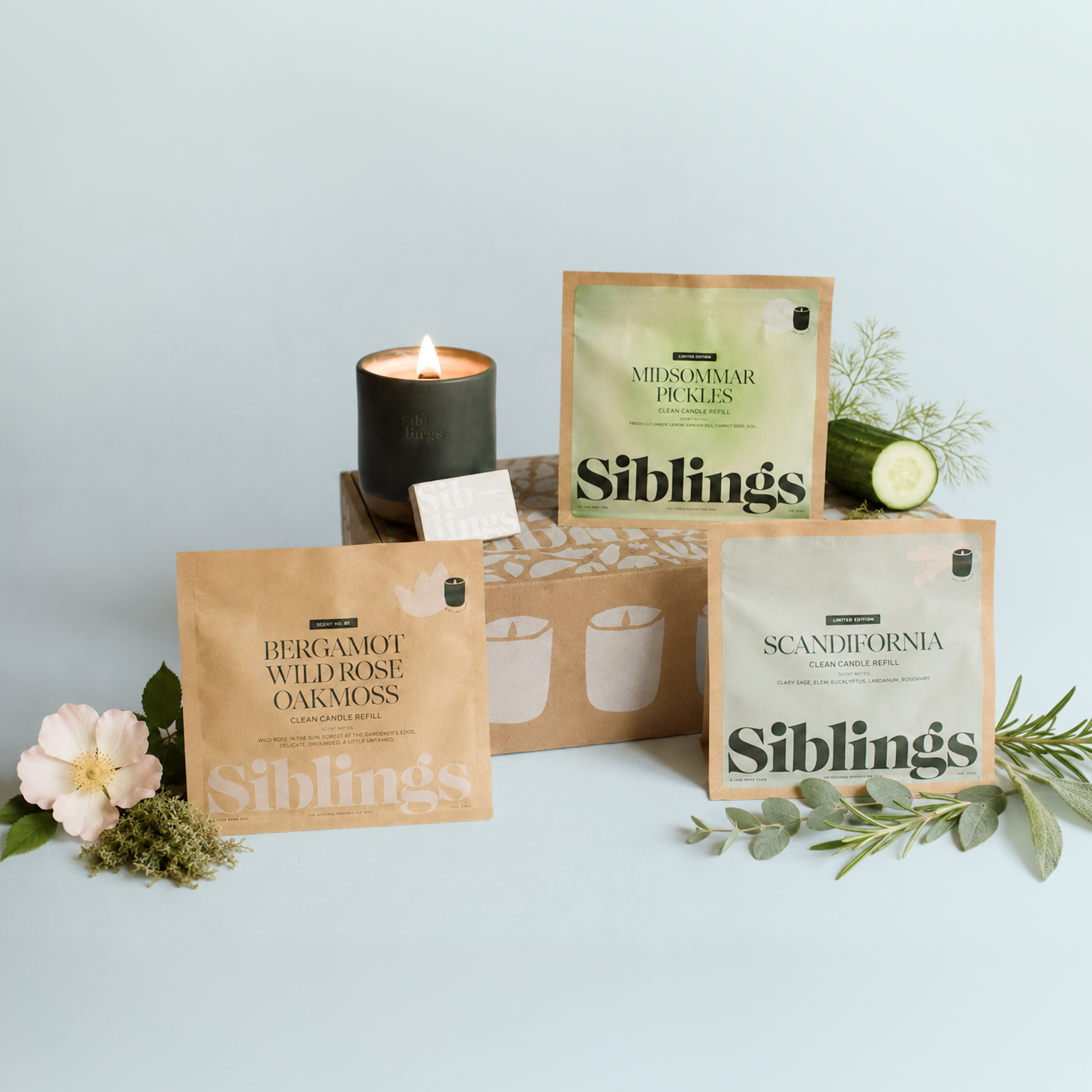 Three 'Siblings' brand candle refill pouches are displayed with a lit candle and corresponding botanical ingredients.
