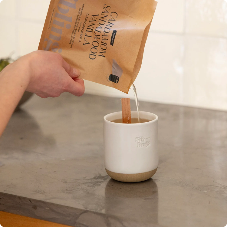 A person pours a candle refill from a paper pouch into a white ceramic vessel with a wood wick.