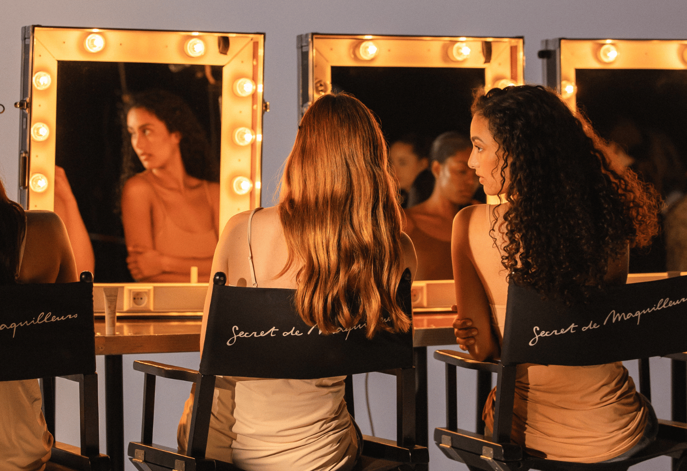 Two women seen from behind, sitting in director's chairs and looking into lit vanity mirrors.