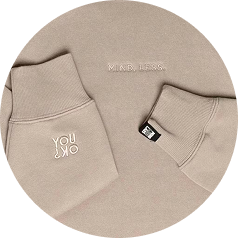 A circular close-up of a beige sweatshirt with embroidered details on the chest and cuffs.