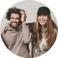 A smiling man with curly hair and a woman in a black cap, both wearing beige hoodies.