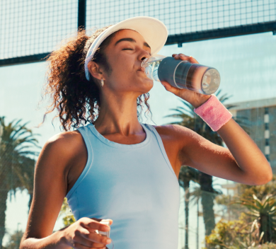 Woman in sports attire drinking water outdoors, with palm trees in the background.