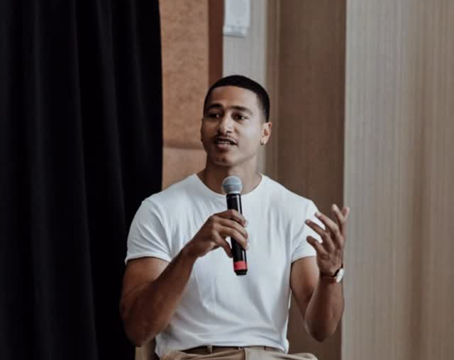 A young man in a white t-shirt holding a microphone speaks and gestures with his other hand.