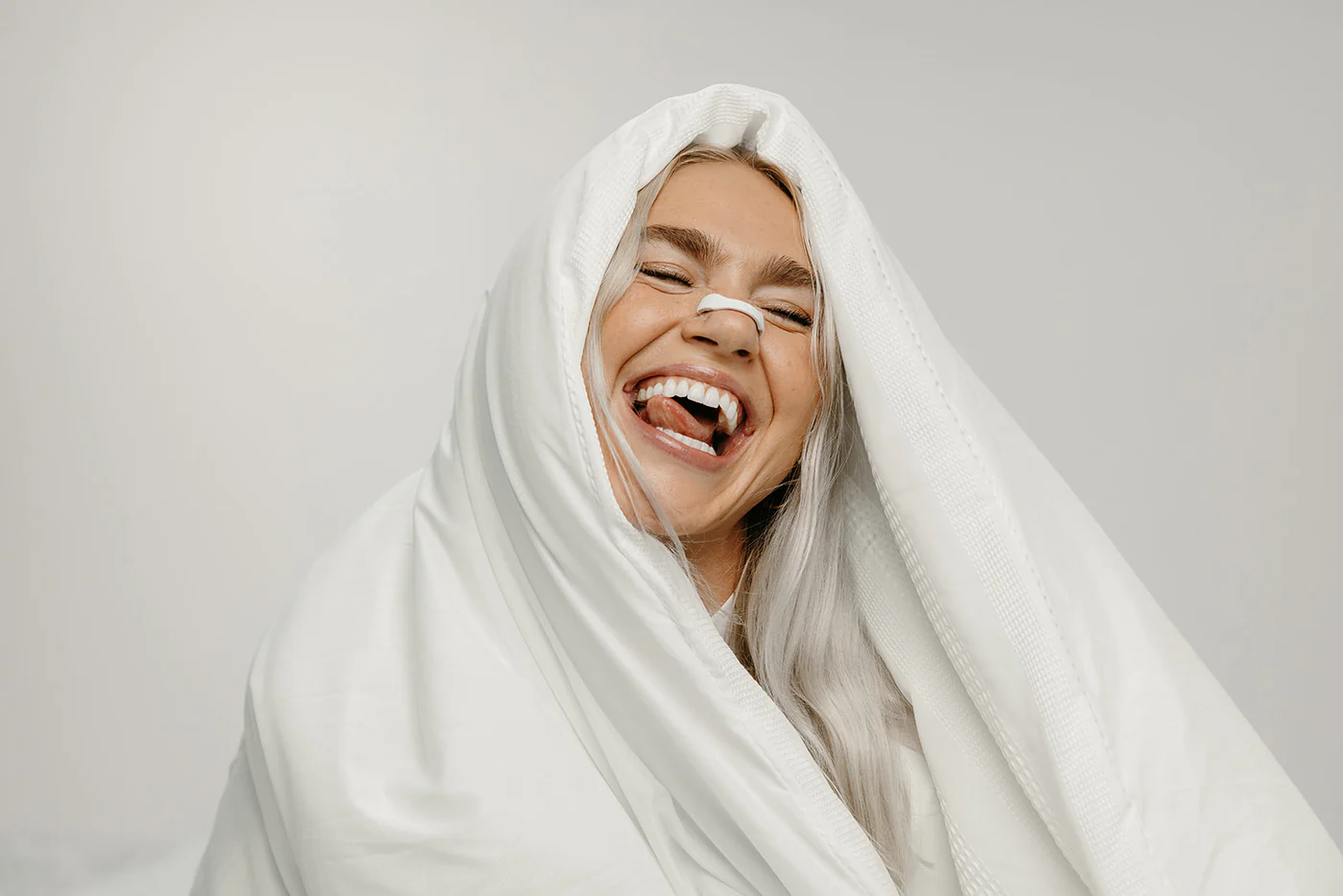 Person laughing, wrapped in a white blanket with a nose strip.