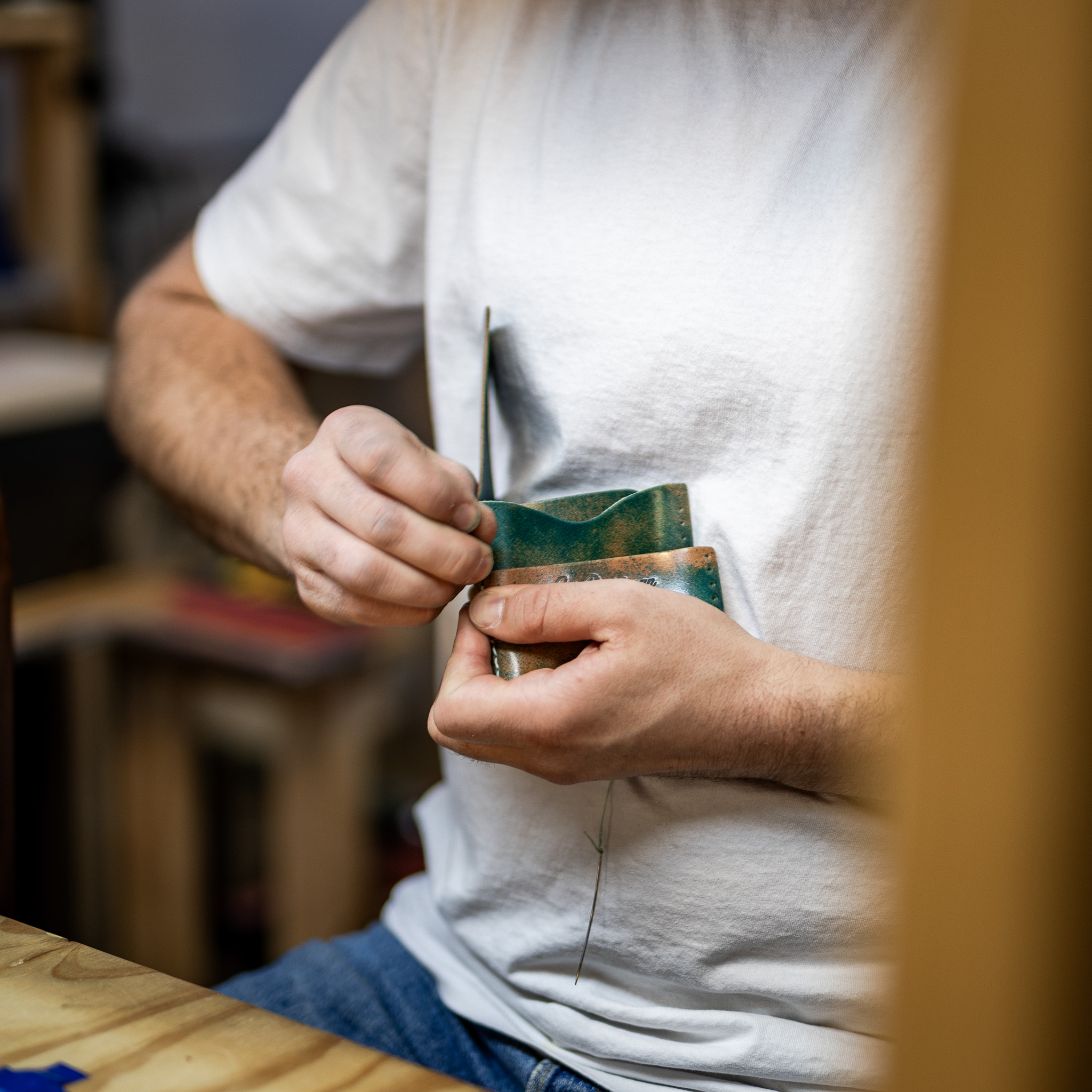 A craftsman in a white t-shirt hand-stitching a small, green and brown leather item.