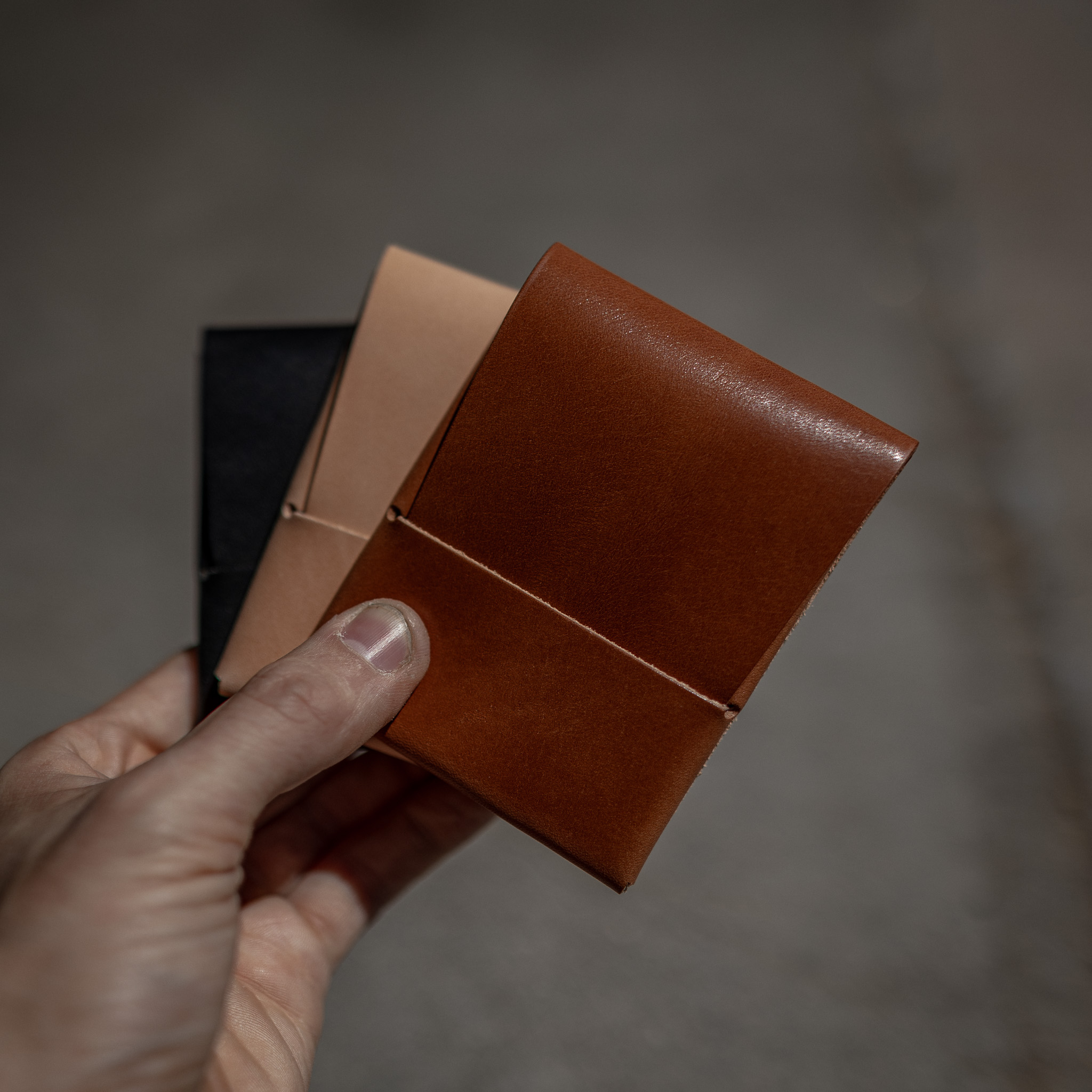 A hand holding three leather cardholders in brown, tan, and black against a gray background.