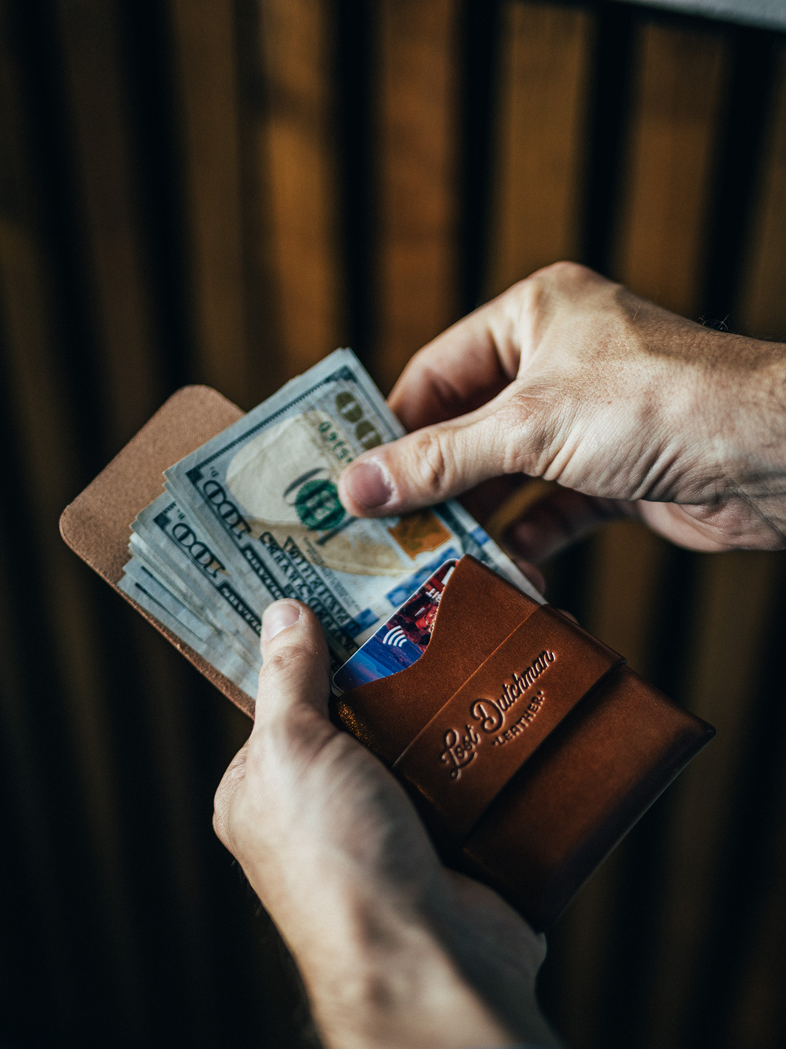 A person's hands putting a stack of hundred-dollar bills into a brown leather wallet.