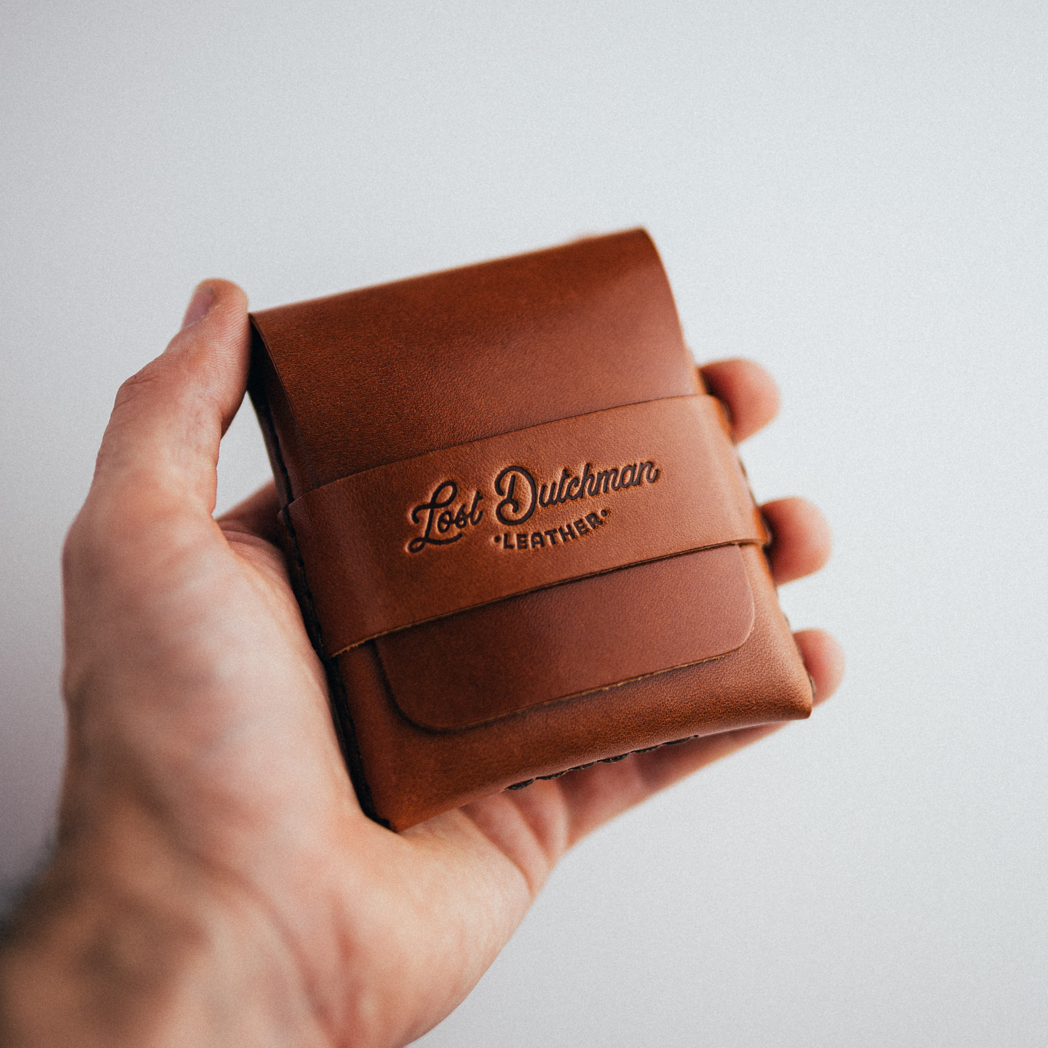 A person's hand holding a brown leather wallet with the Lost Dutchman Leather logo embossed on it.