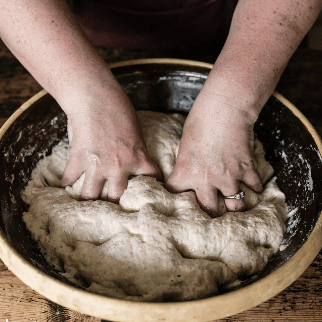 Hands kneading dough in a large bowl on a wooden table.