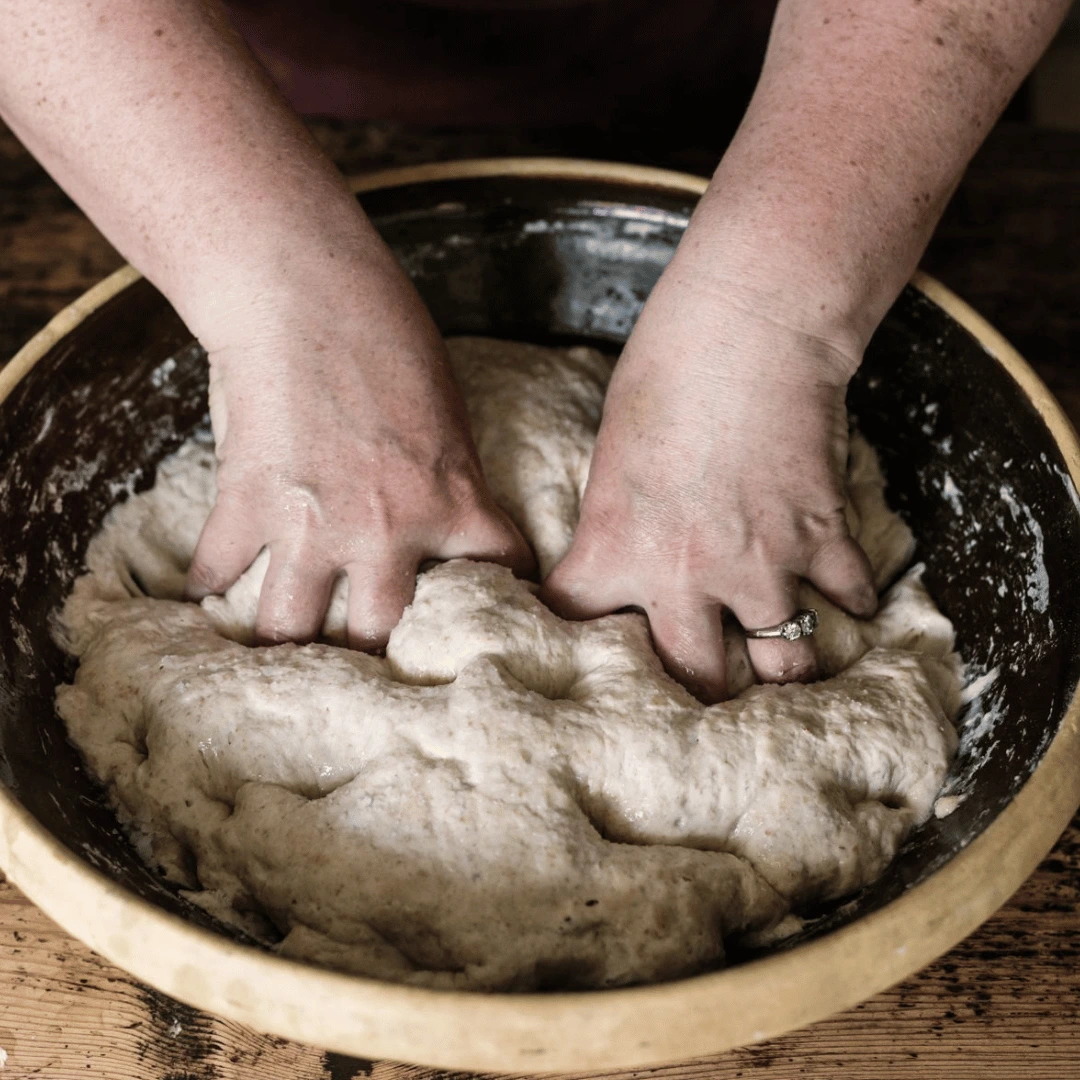 Hands kneading dough in a large bowl on a wooden table.