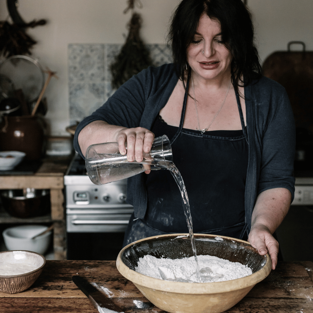 Person pouring water into a bowl of flour in a kitchen setting.