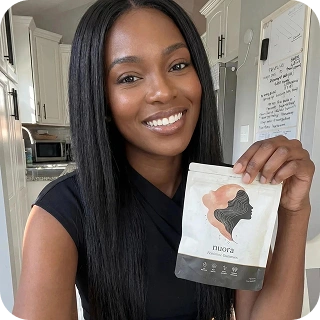 A smiling woman with long, straight black hair holds up a white product pouch in a kitchen.