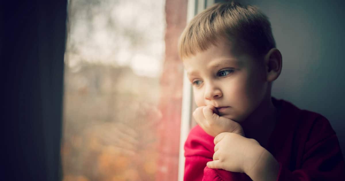Young child in red shirt looking out a window thoughtfully.