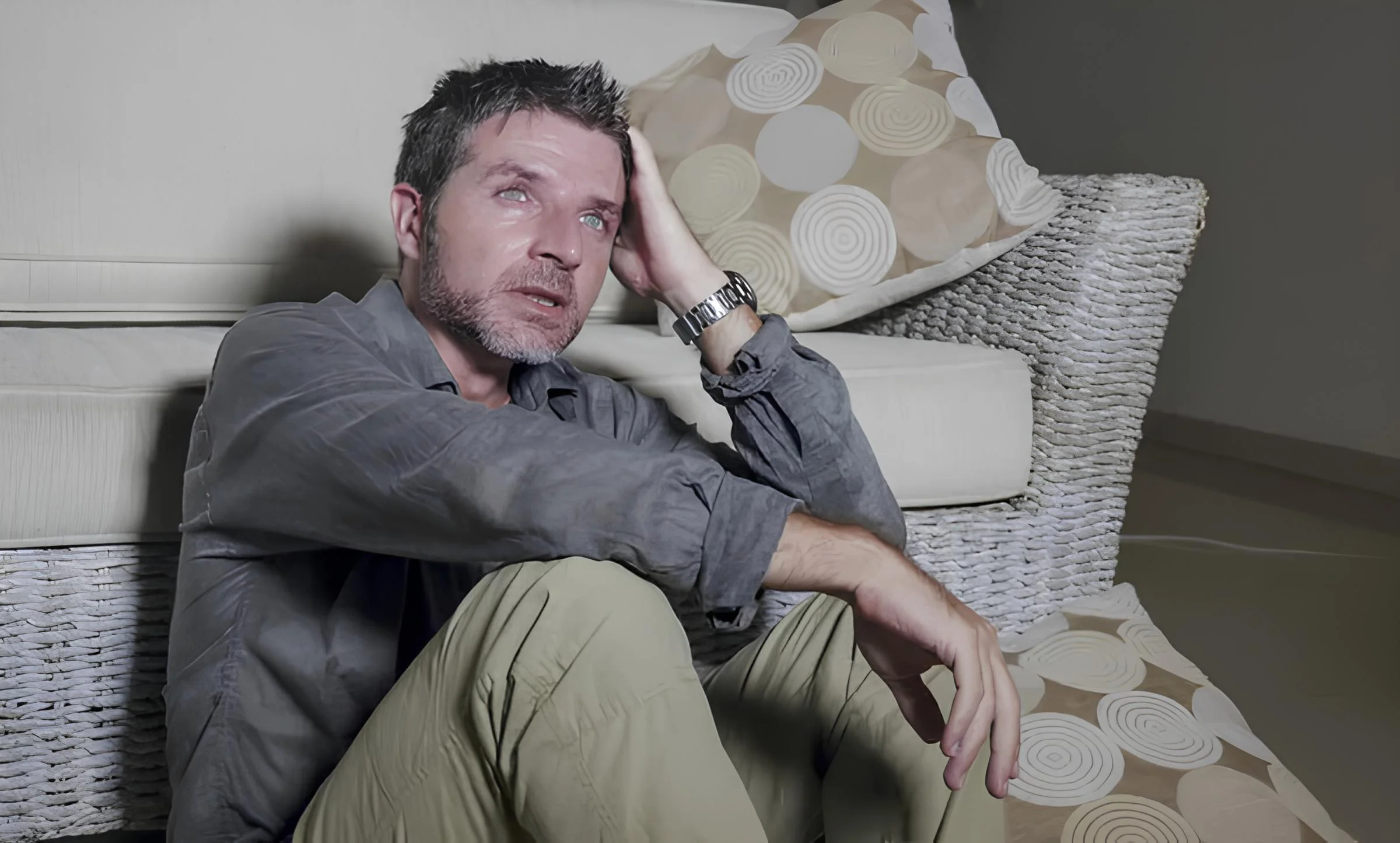 Man sitting on the floor, leaning against a couch, looking contemplative.