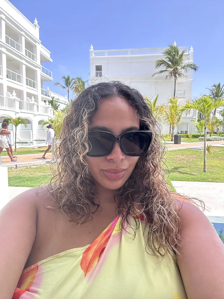 A woman in sunglasses takes a selfie outdoors with a white building and palm trees in the background.