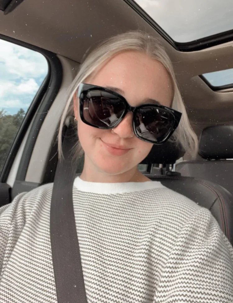 A blonde woman wearing large sunglasses and a striped sweater smiles for a selfie inside a car.