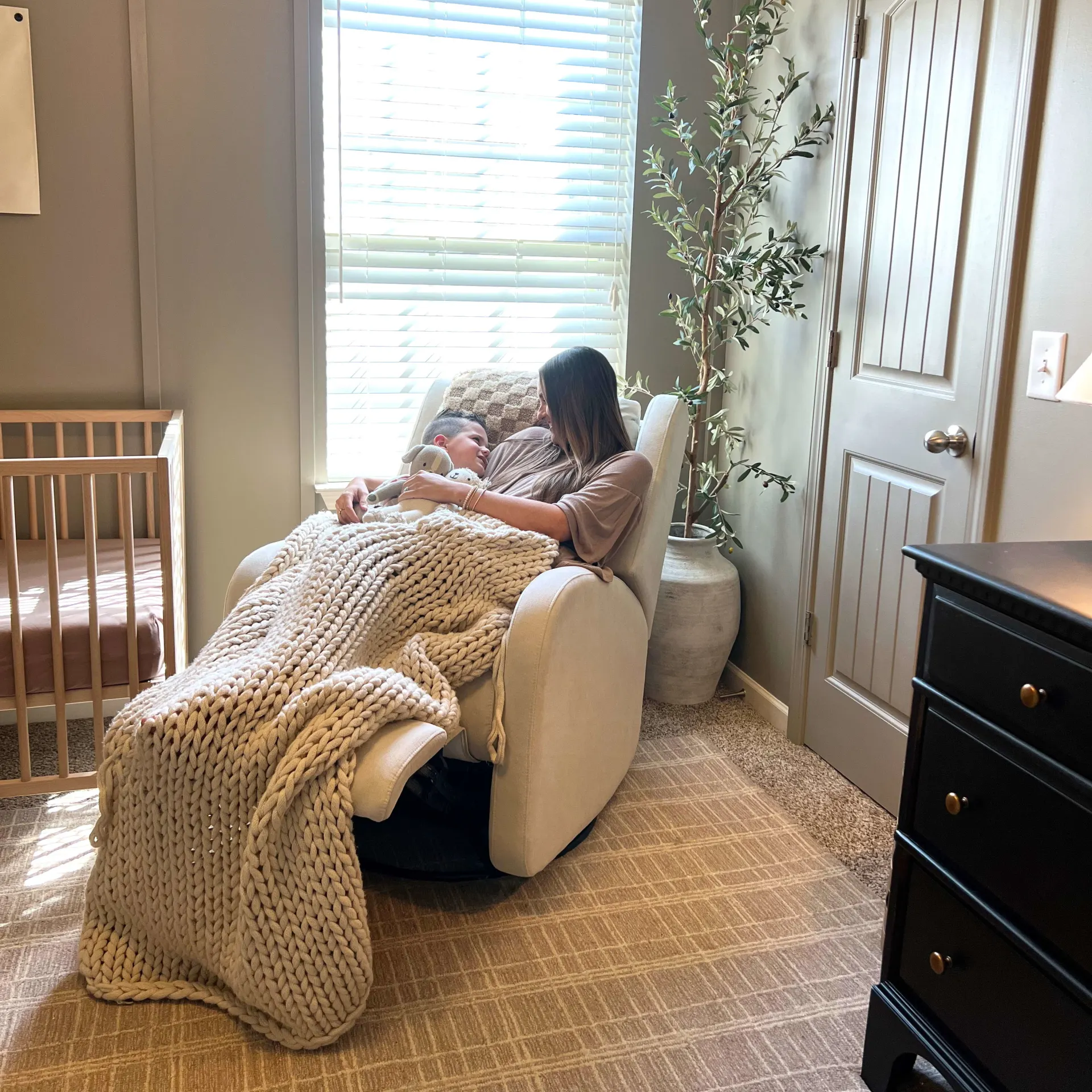 A woman and child cuddle in a rocking chair in a nursery, covered by a large knit blanket.