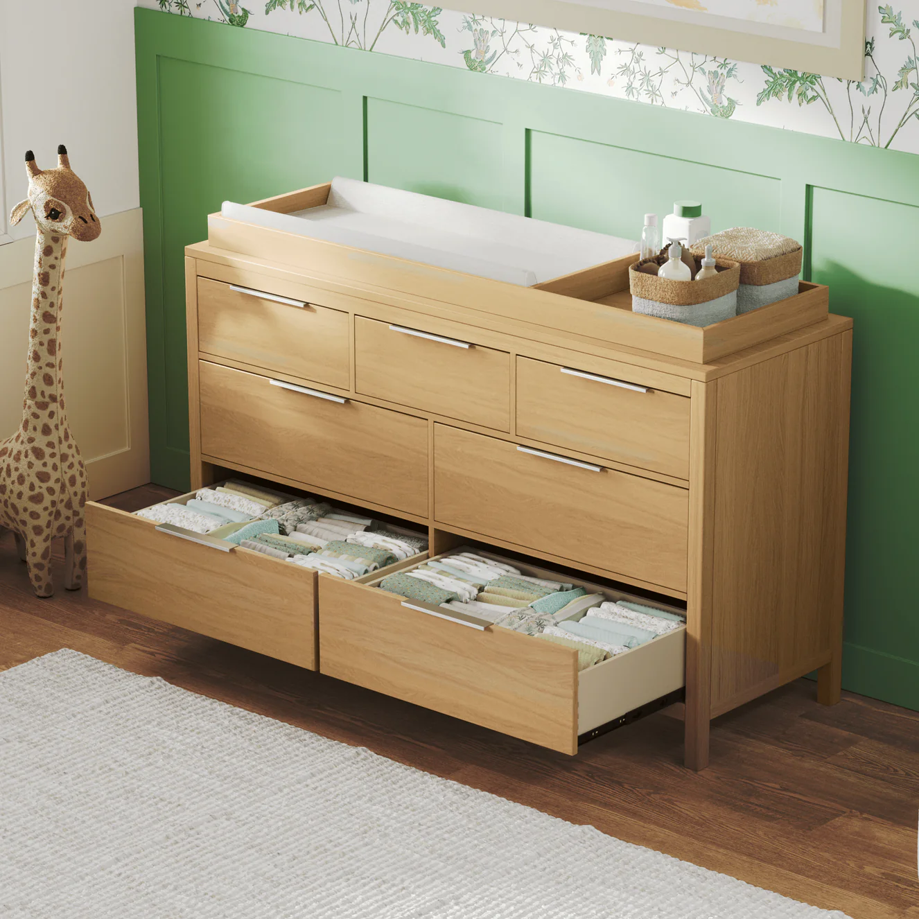 A light wood changing table dresser with open drawers in a nursery next to a large stuffed giraffe.