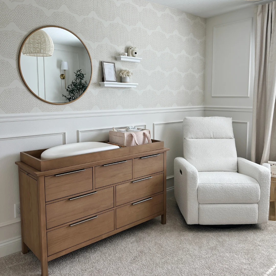 A nursery with a wooden changing table, a white armchair, and a round mirror on the wall.