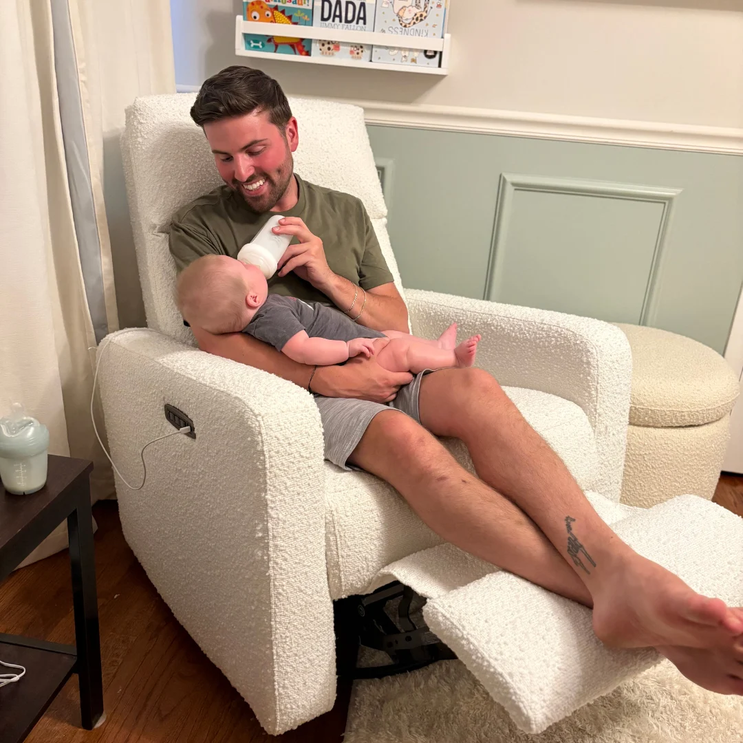 A smiling man sits in a white recliner chair, bottle-feeding a baby he is holding in his arms.