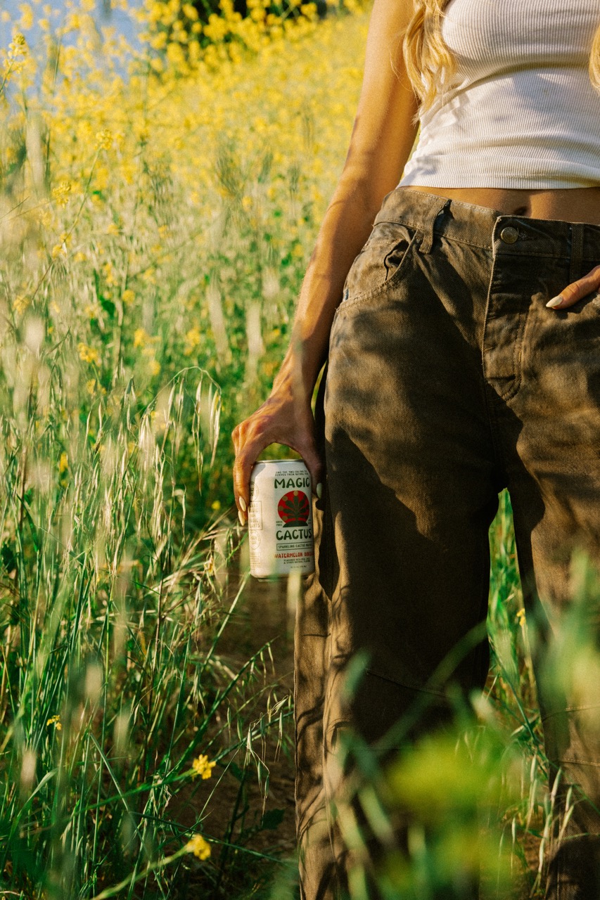 A person in brown pants holds a can of Magic Cactus in a field of yellow flowers.