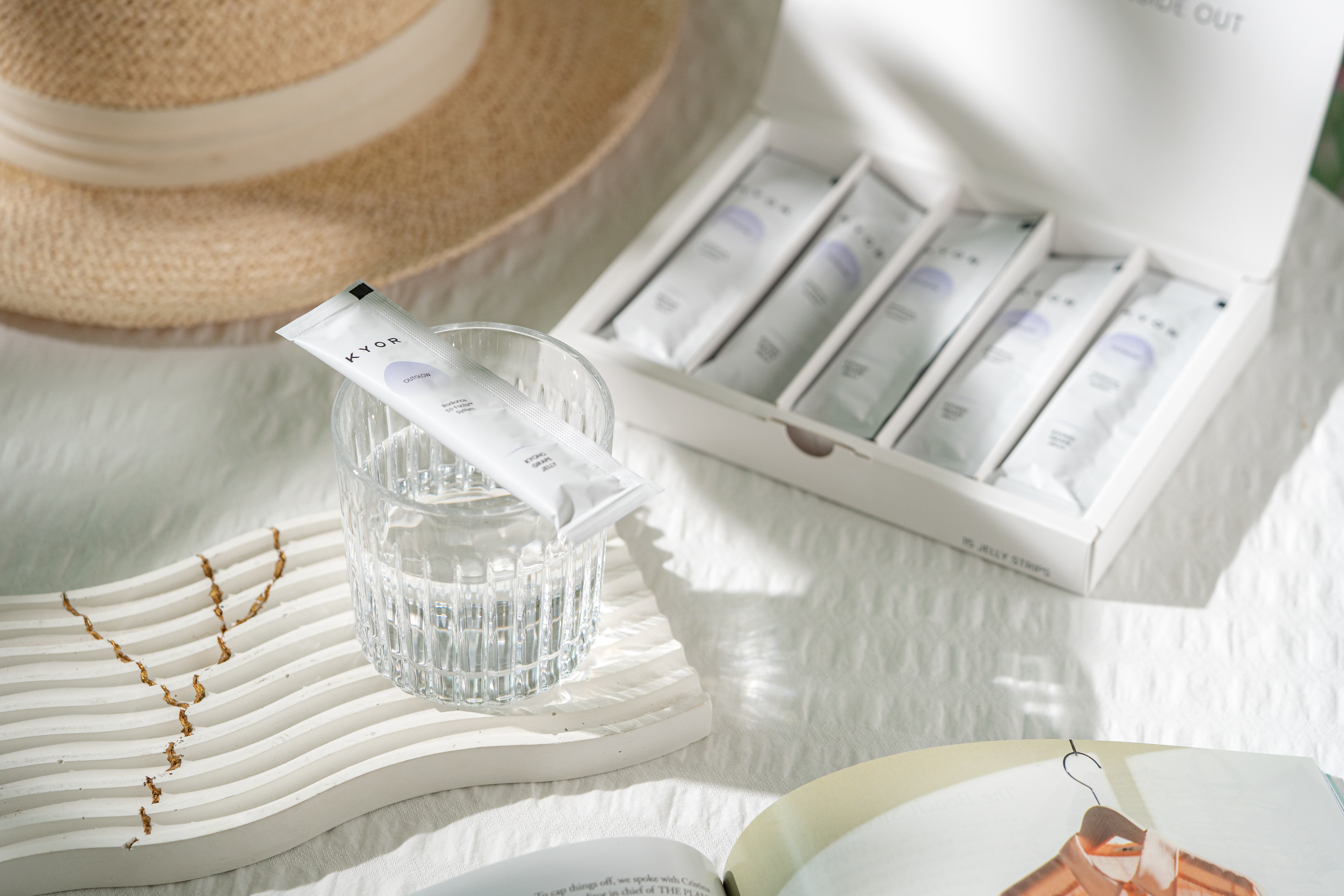 A KYOR sachet rests on a glass of water next to an open box of the product and a hat.