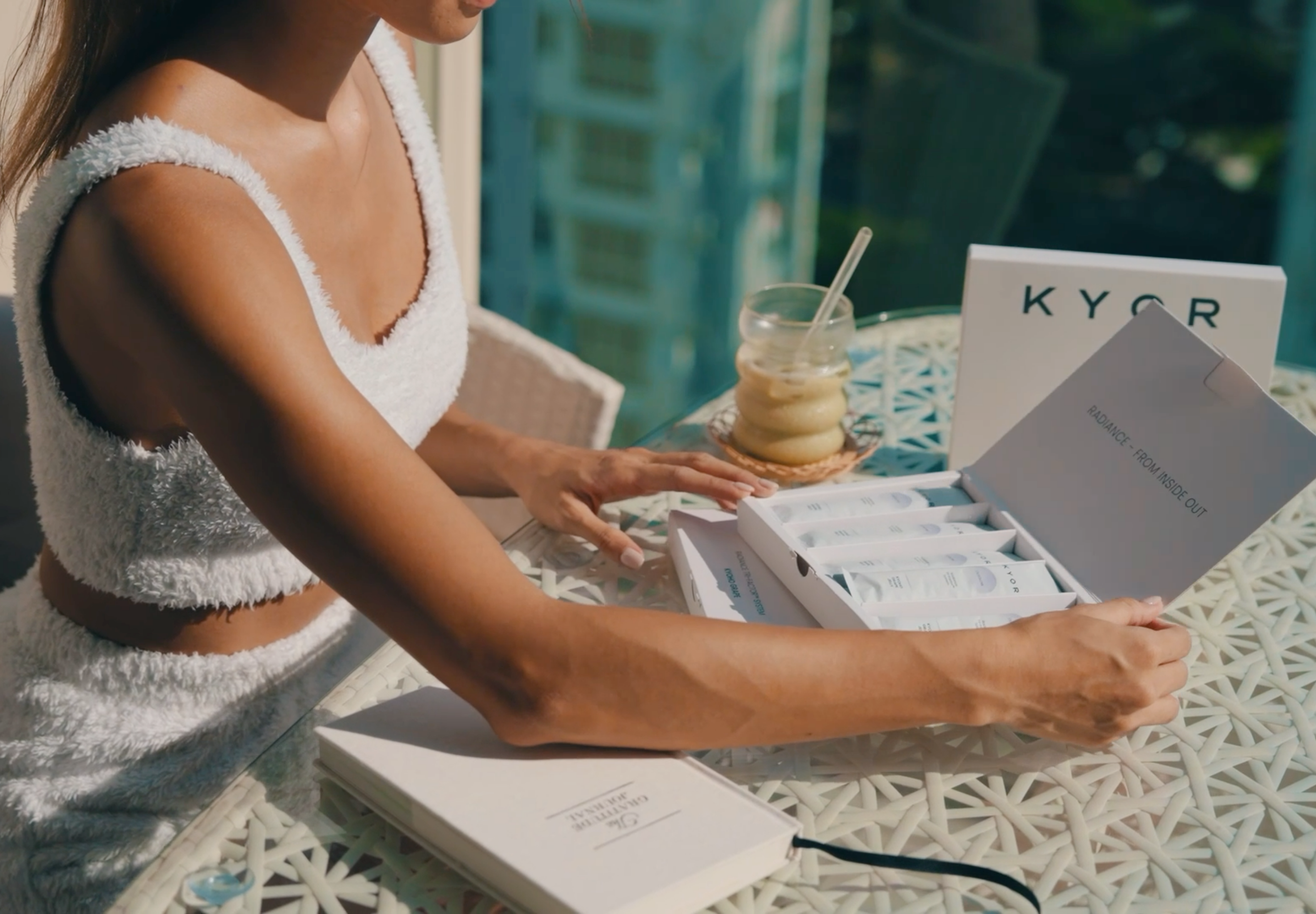 A woman in a white outfit opens a box of KYOR beauty products at a woven table.