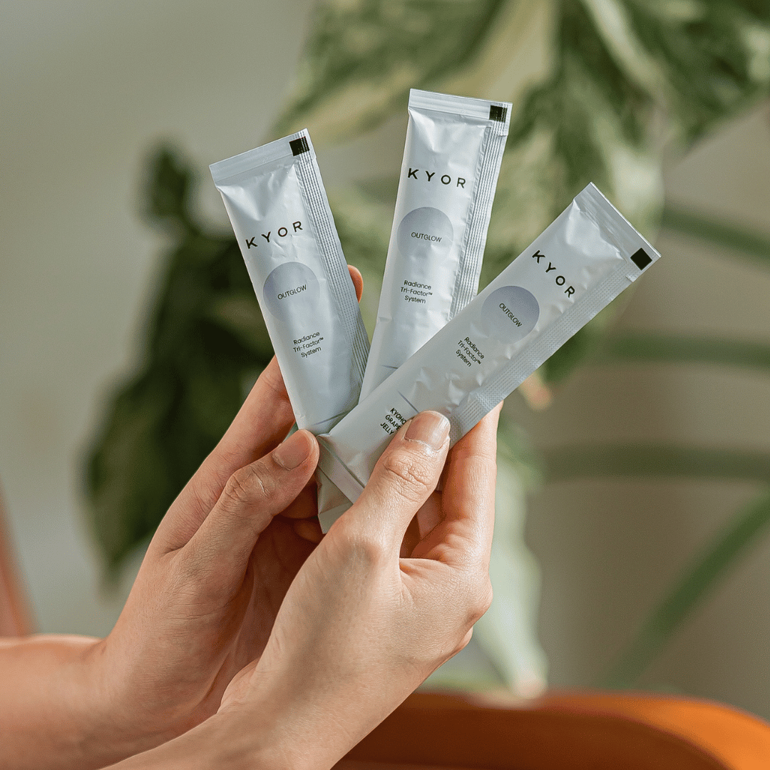 A pair of hands holding three silver packets of KYOR OUTGLOW supplement against a leafy background.