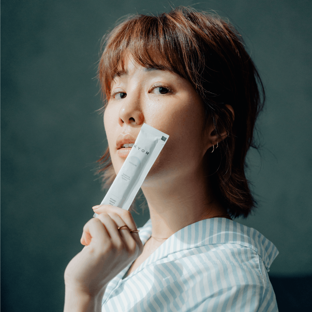 A woman with reddish-brown hair in a striped shirt holds a white KYOR sachet to her lips.