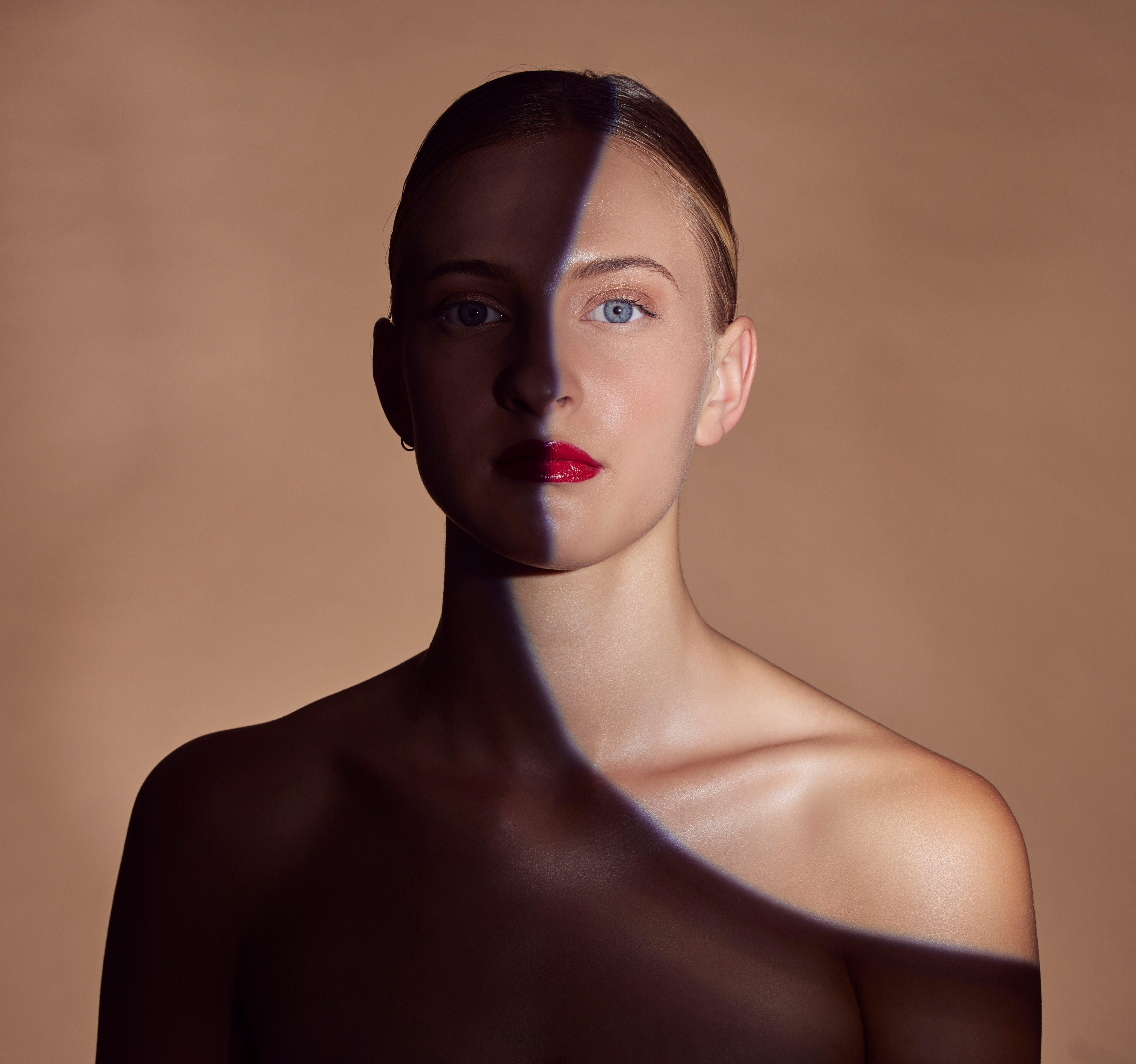 A woman's face and bare shoulders with a diagonal shadow cast across them, against a plain brown background.
