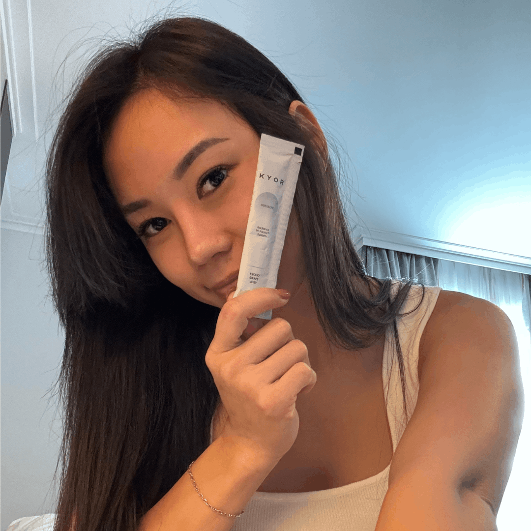 A woman with long dark hair holds up a white product sachet with the word KYOR on it.