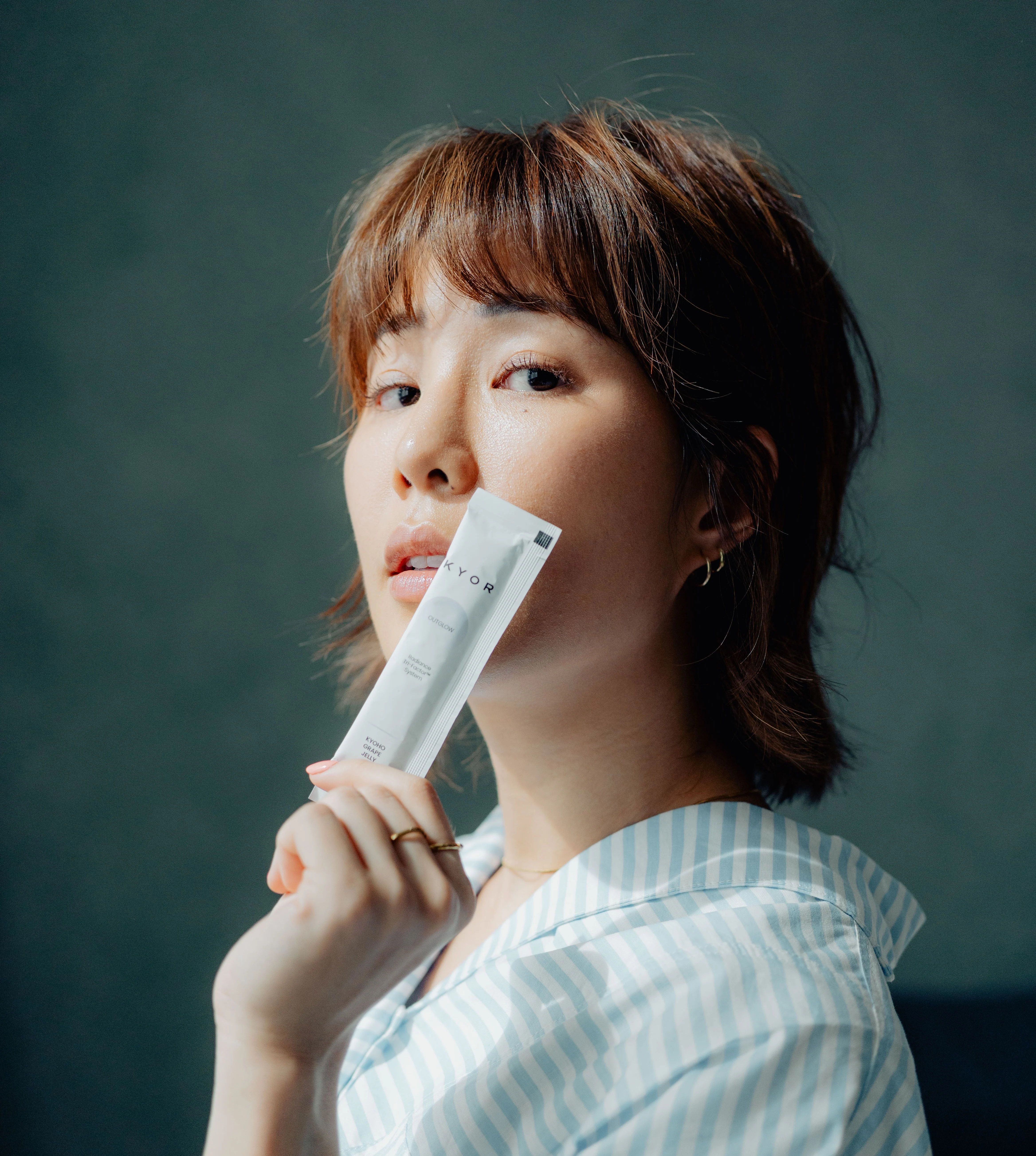 A woman with short brown hair holds a white packet in front of her mouth, looking at the camera.