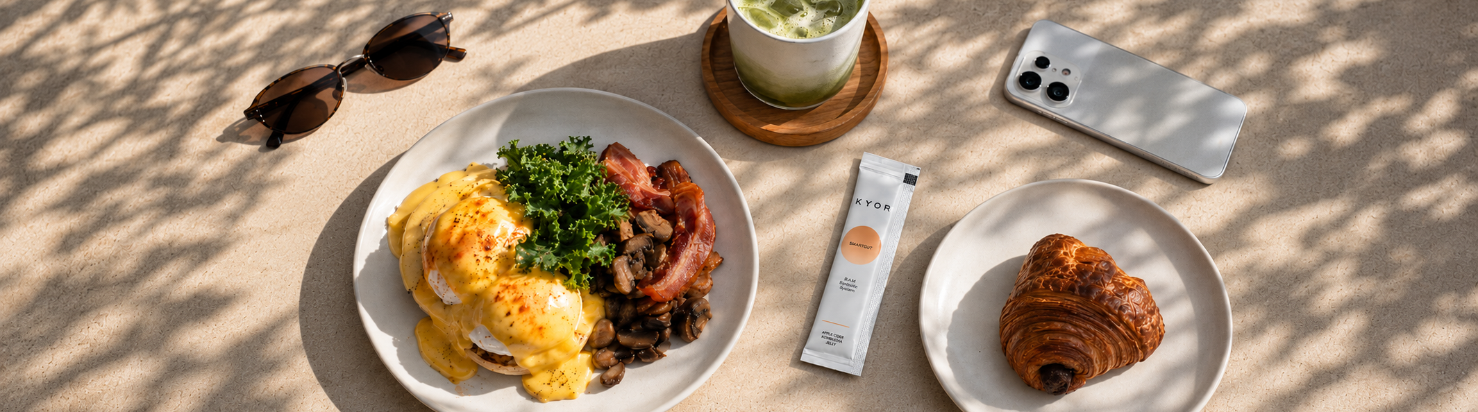 An overhead shot of a brunch spread with eggs Benedict, a croissant, a matcha latte, and sunglasses.