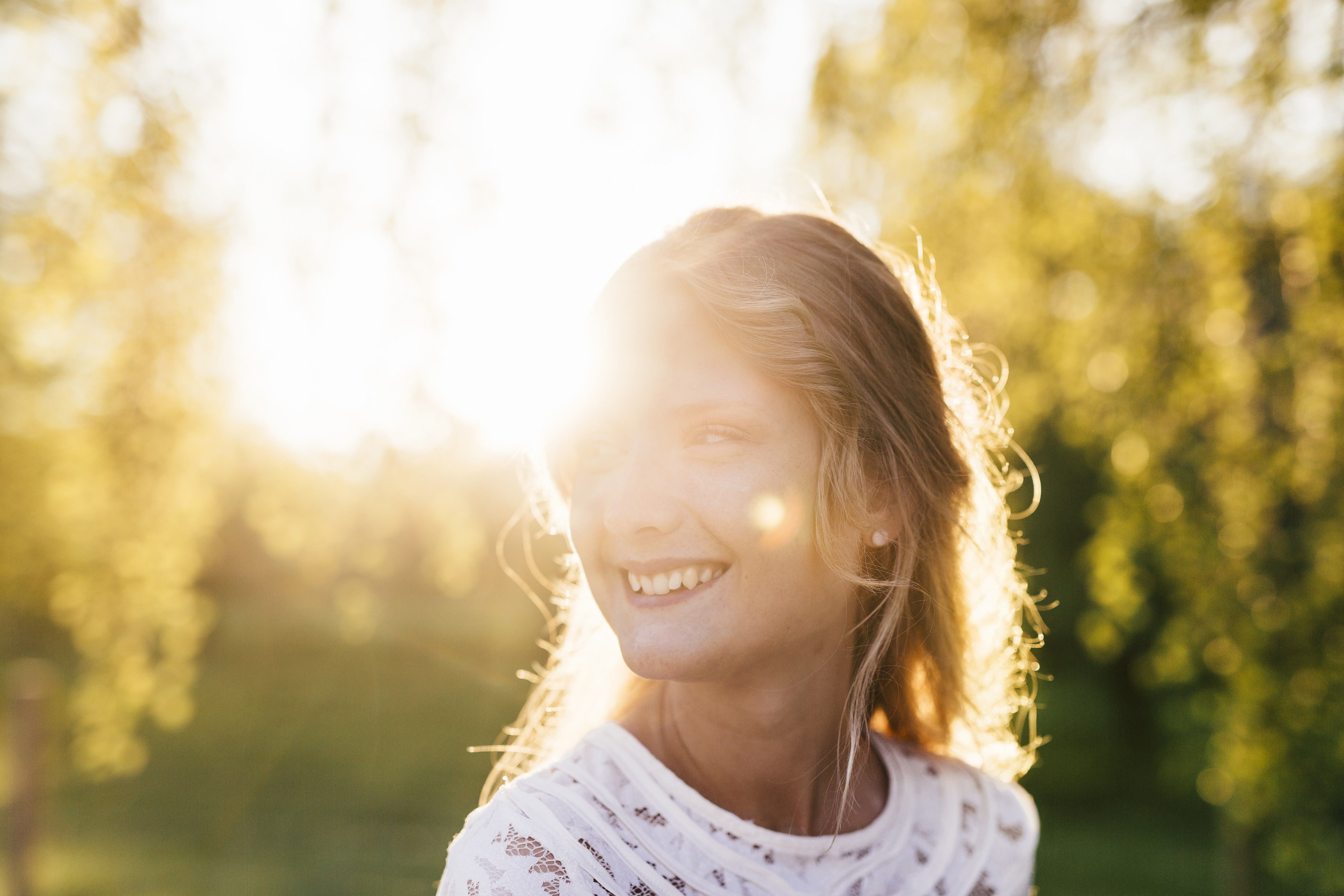 A smiling woman with blonde hair, backlit by the sun in a grassy, wooded area.