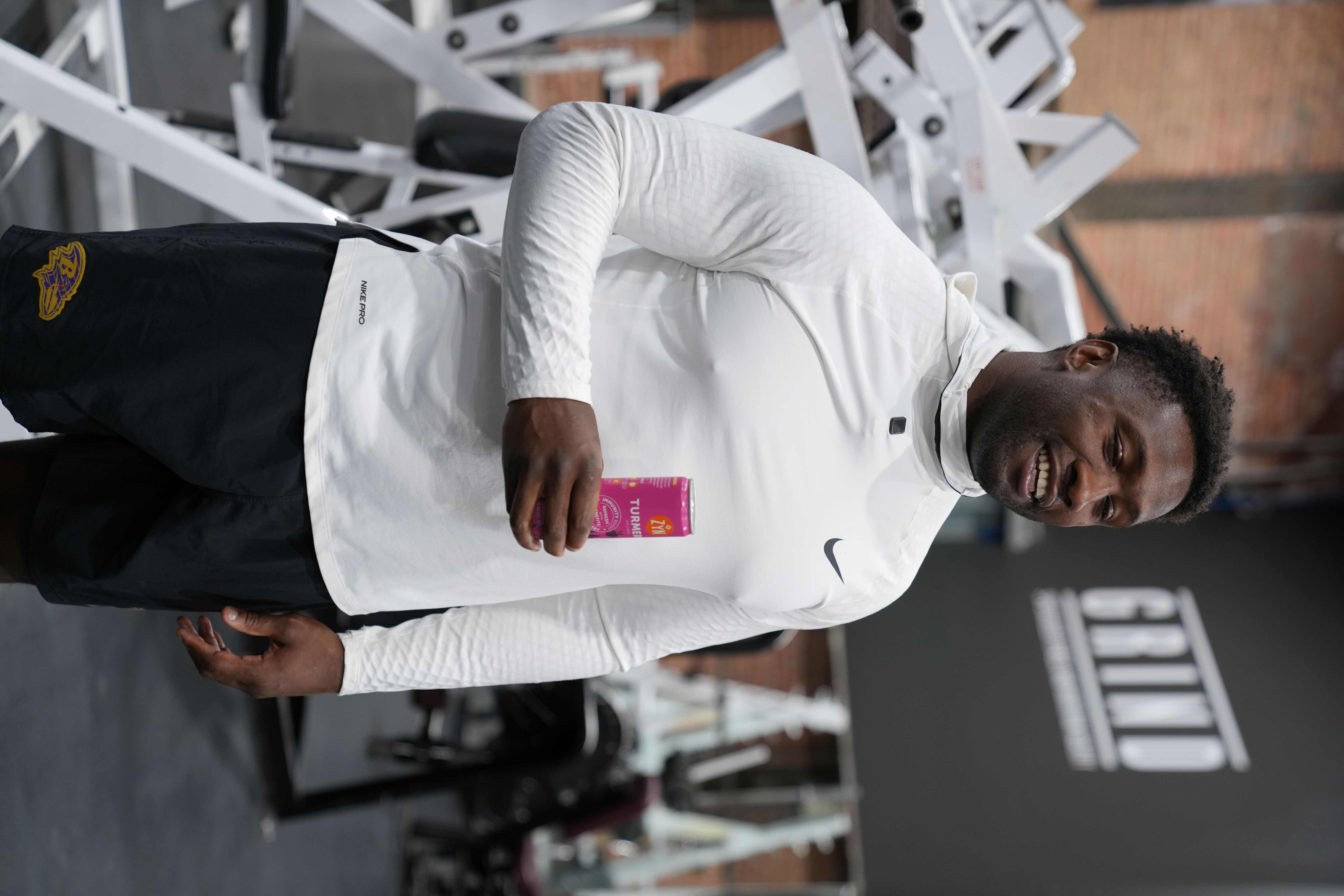 Man in gym holding a pink can, wearing white top and black shorts.