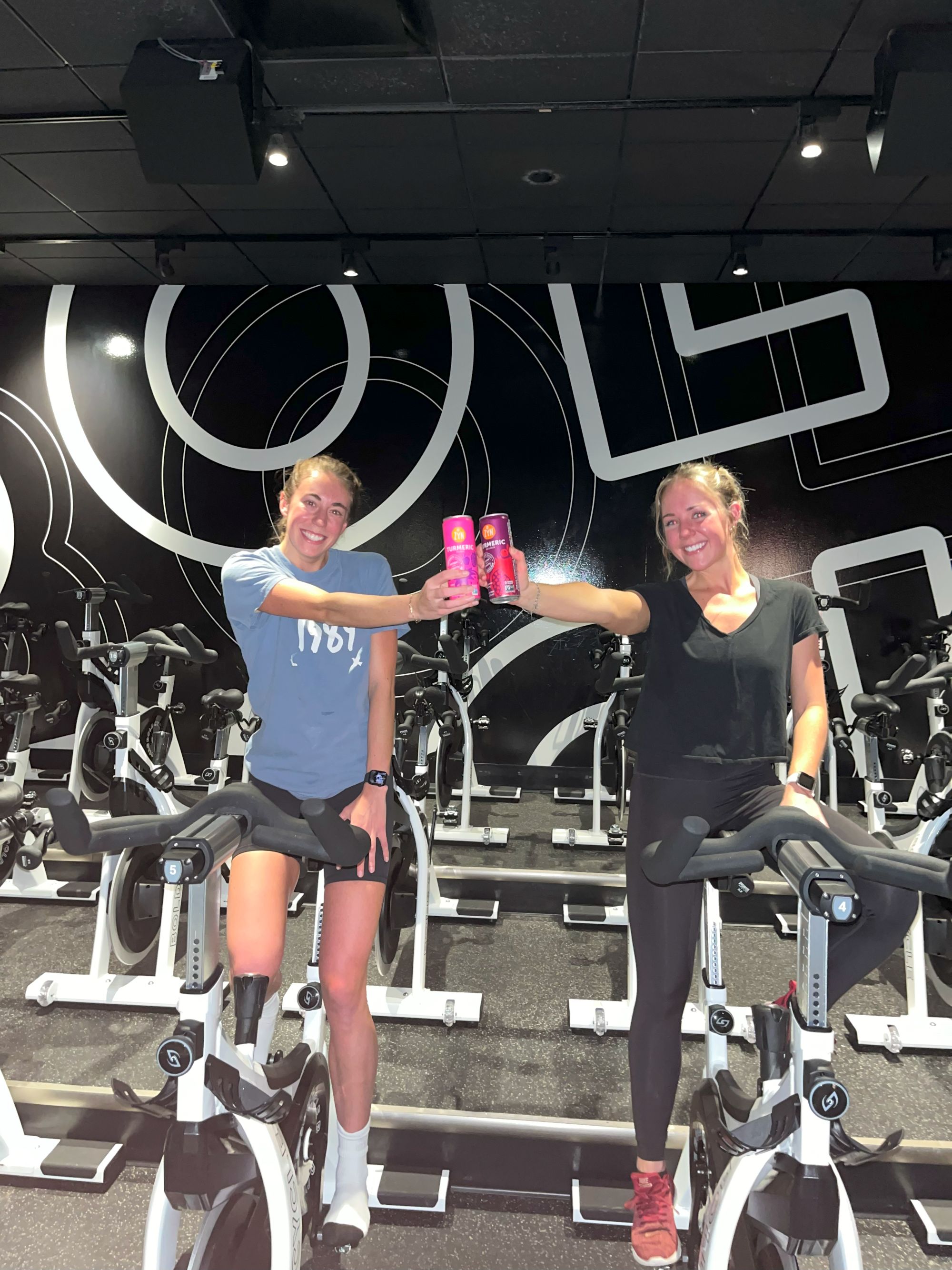 Two women on stationary bikes holding energy drinks in a gym.