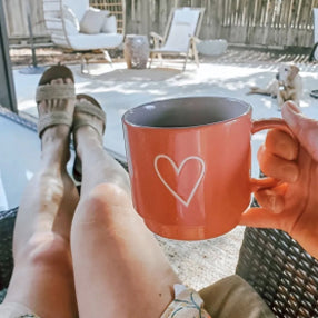 Person relaxing with a heart-decorated mug, feet up, outdoors.