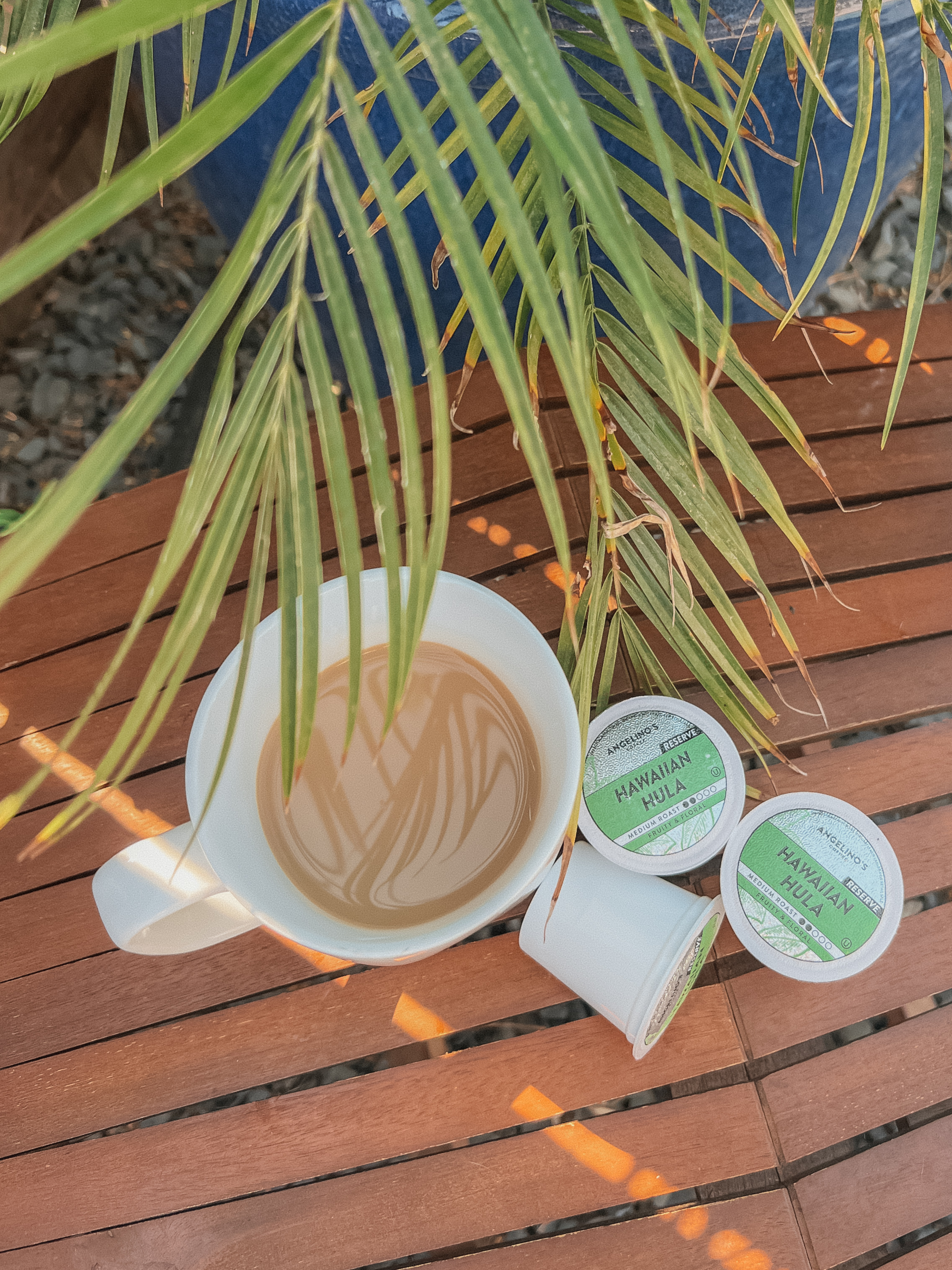 Cup of coffee and Hawaiian Hula pods on a wooden table with palm leaves.
