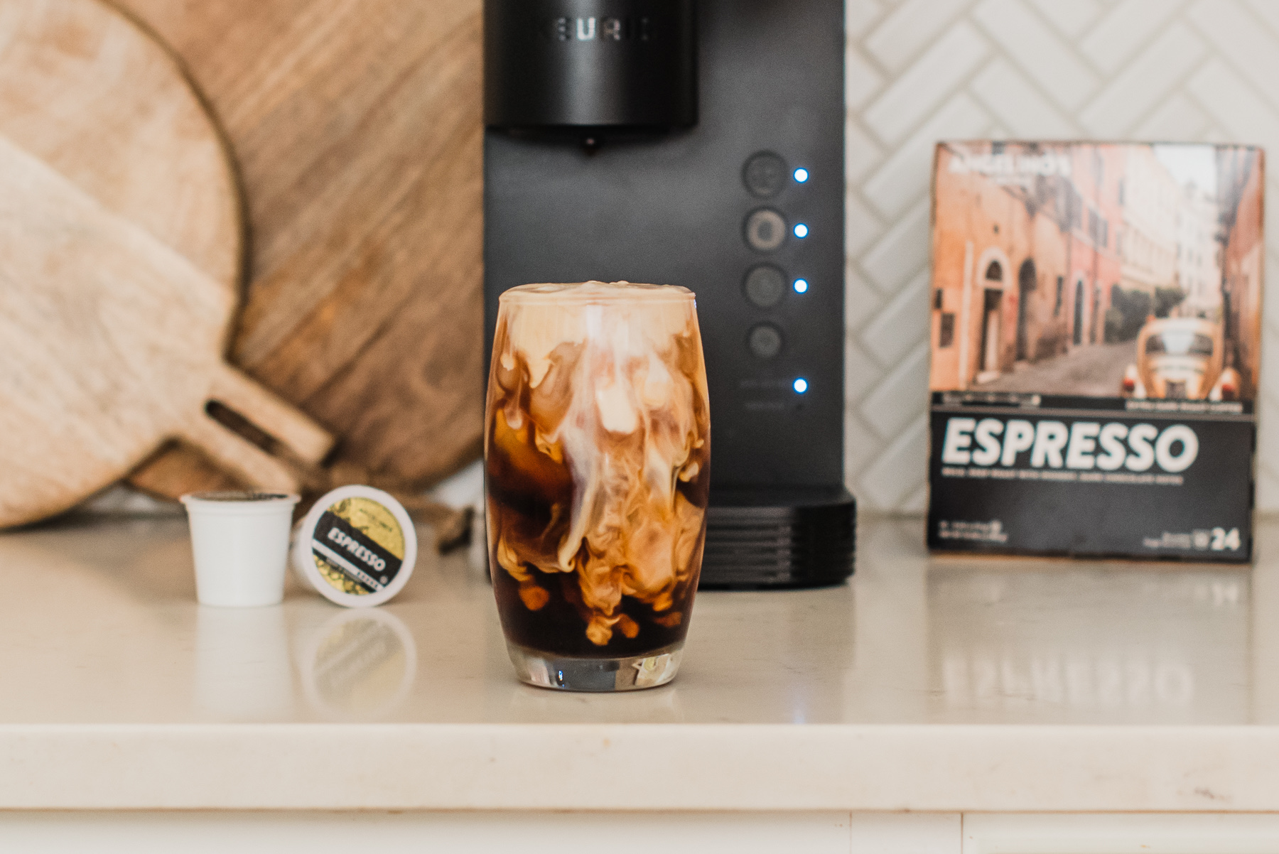 Iced coffee with milk swirls, espresso pods, and a coffee machine on a kitchen counter.