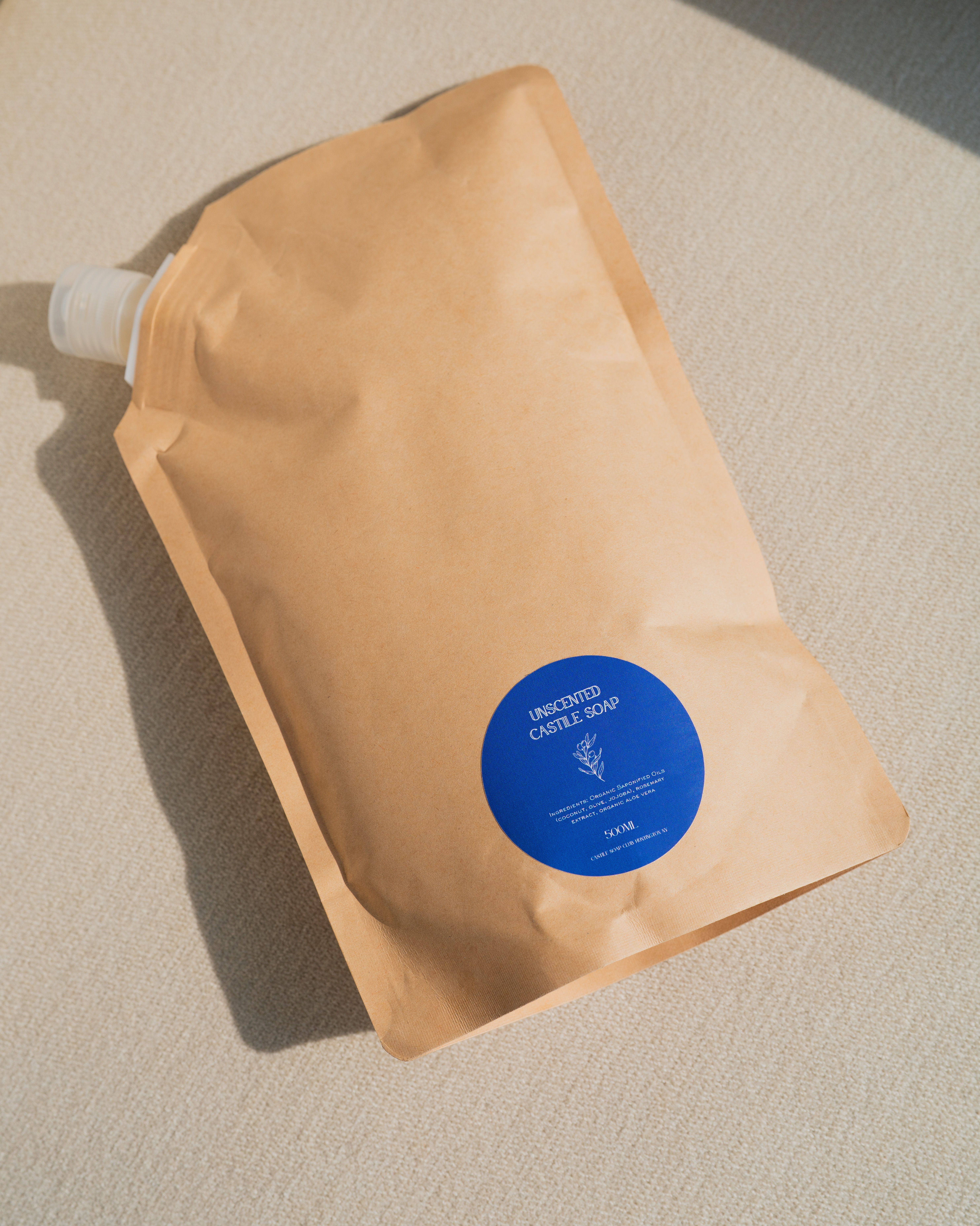A brown paper refill pouch of unscented castile soap with a round blue label on a textured surface.