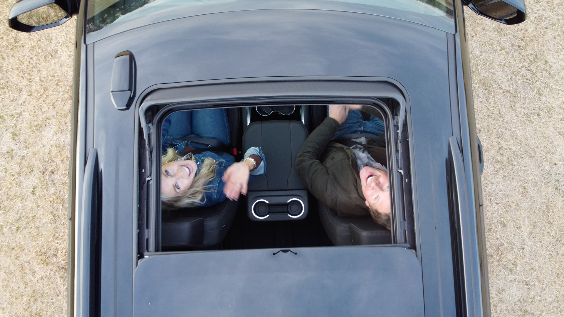An aerial view of a man and woman smiling and looking up through a car's open sunroof.