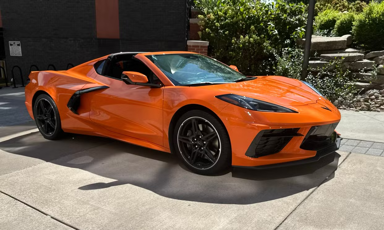 A bright orange Corvette C8 sports car is parked on a paved driveway in the sunlight.