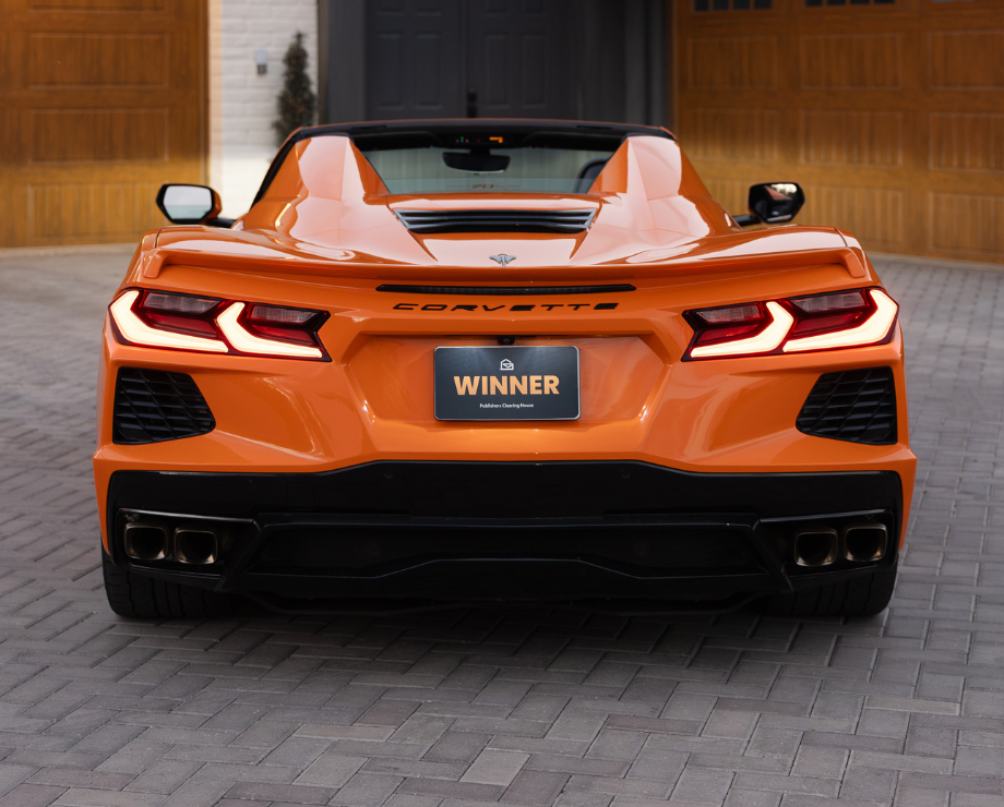 A bright orange Chevrolet Corvette convertible parked on a paved driveway in front of a garage.