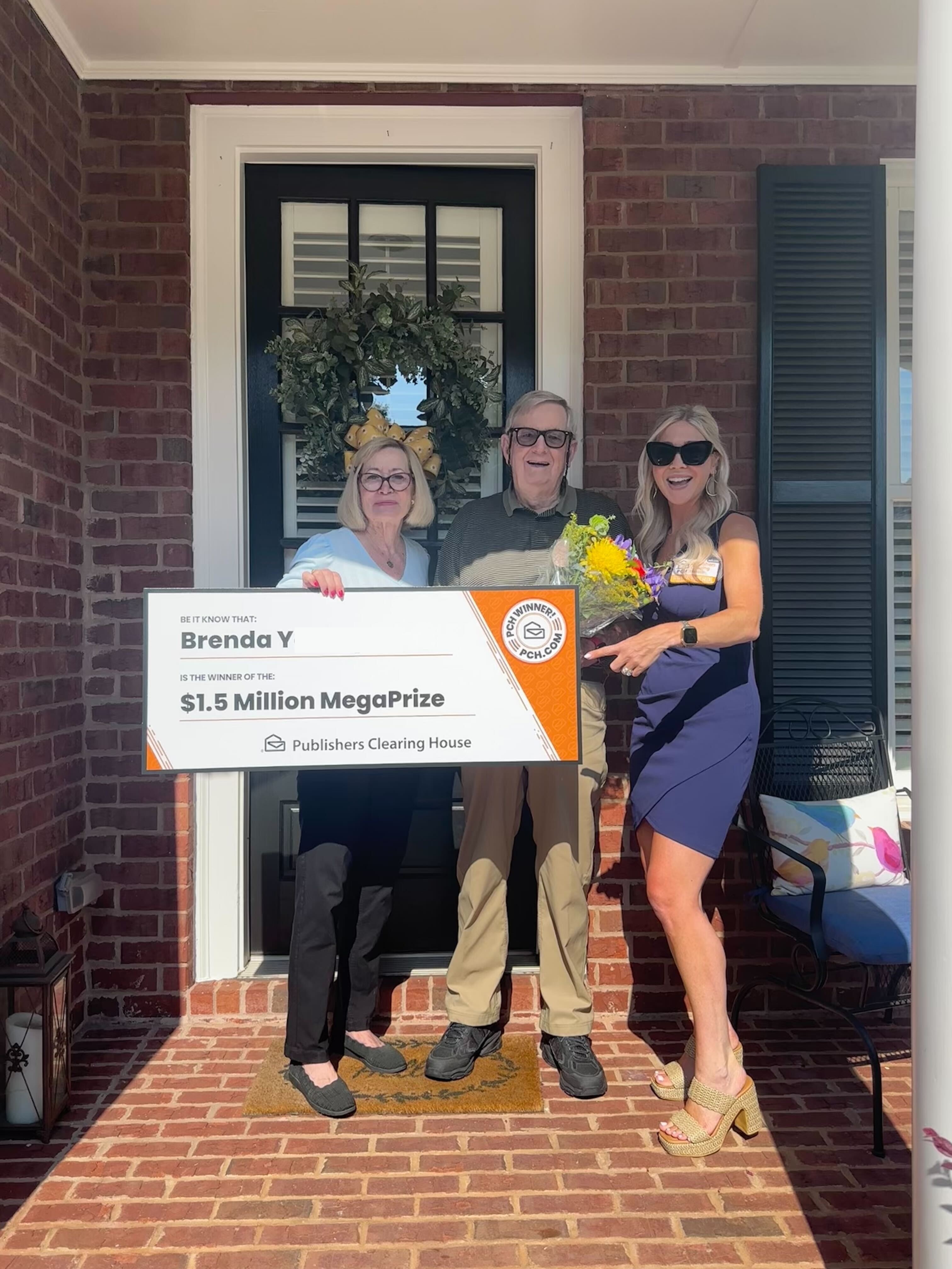 An older couple and a representative hold a large Publishers Clearing House check for $1.5 million on a porch.
