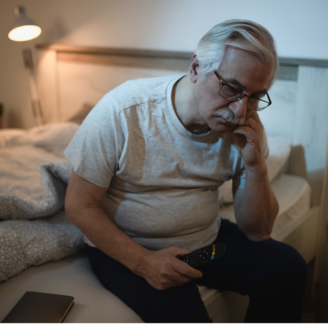 Elderly man sitting on bed, holding a remote, looking thoughtful.