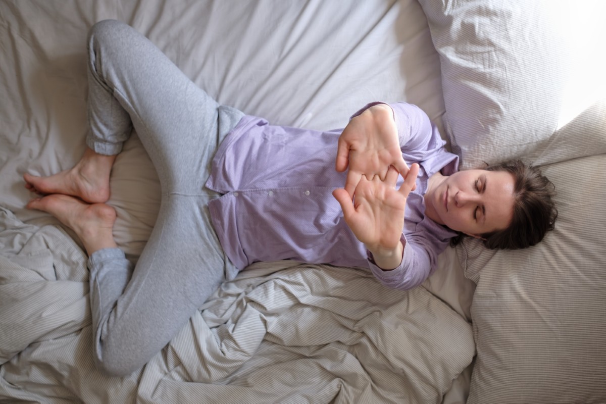 Person lying on bed with arms stretched and crossed, wearing grey pants and purple shirt.