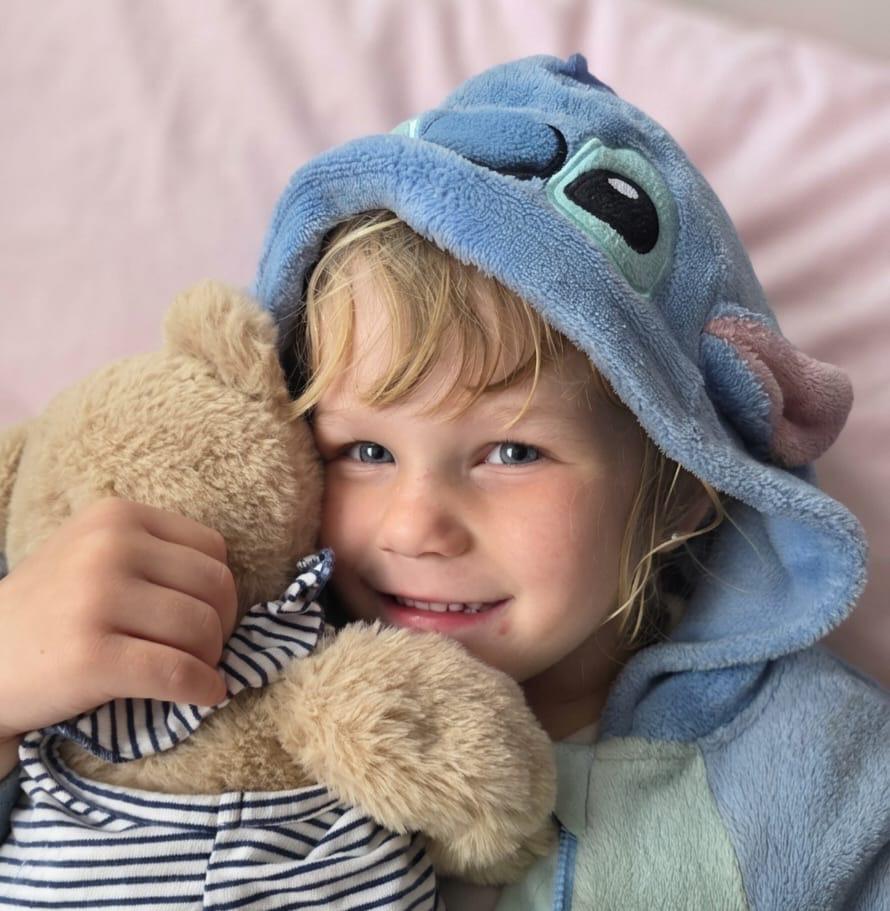 A smiling child in a blue character hoodie hugs a teddy bear.