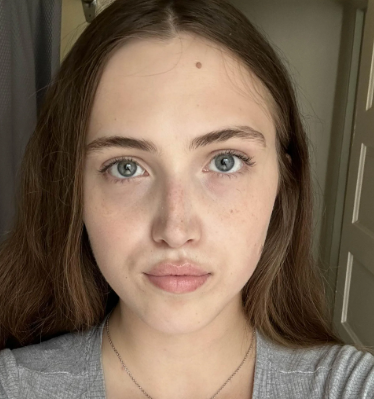 A close-up selfie of a young woman with long brown hair and blue eyes looking at the camera.