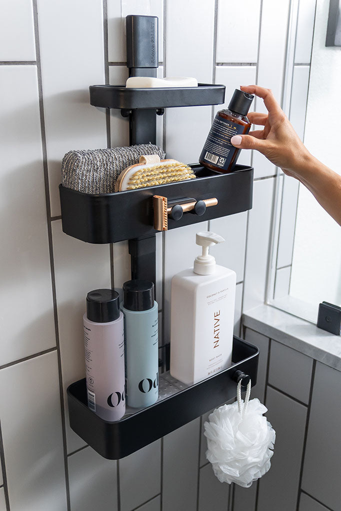 A hand places a bottle of soap into a black, multi-level shower caddy against a white tiled wall.